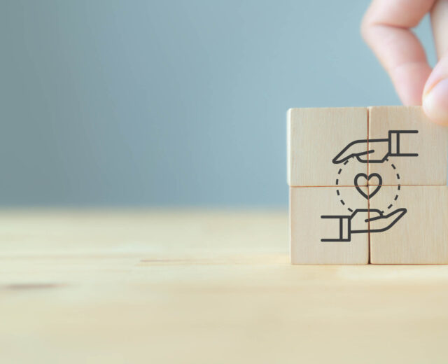 Four wooden blocks with a hand, heart, and hand design are arranged to form a complete image. A hand is placing the final block.