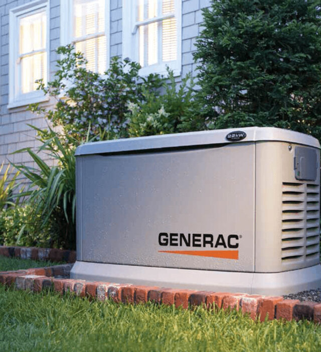 A Generac generator installed outside a house on a brick platform, surrounded by grass and small shrubs.