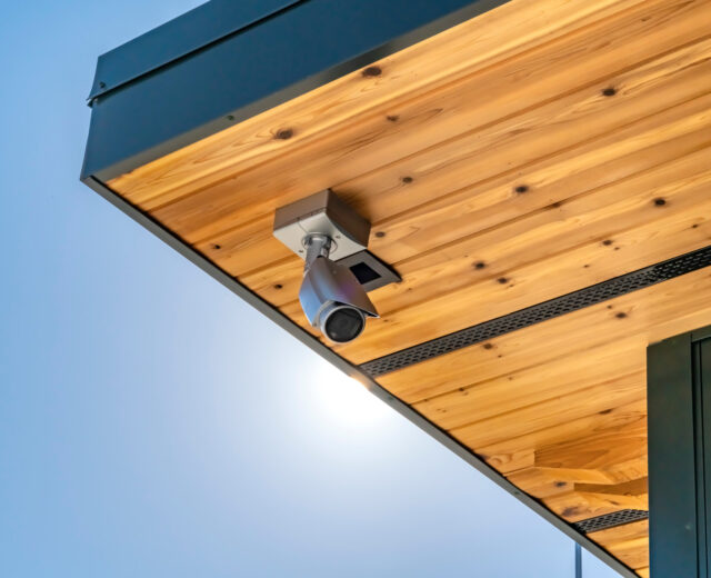Surveillance camera mounted on a wooden ceiling under a clear blue sky.