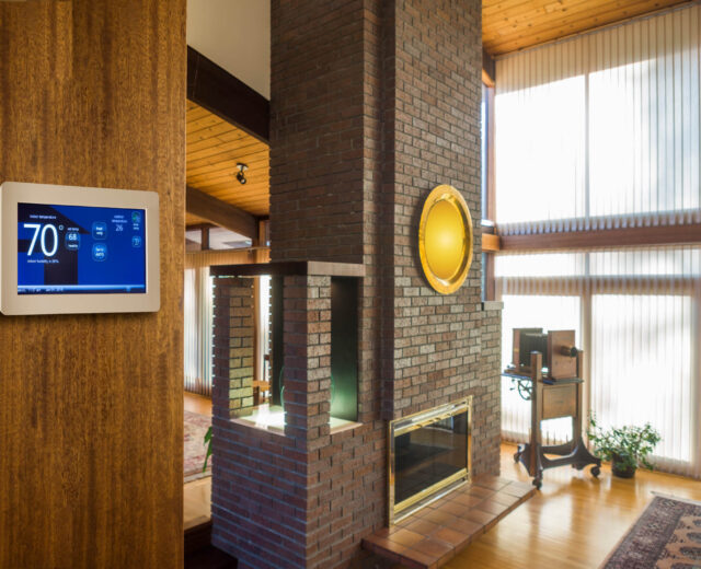 A modern living room with a brick fireplace, large windows, and a smart thermostat on the wall displaying 70 degrees.