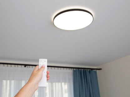A hand holds a remote control pointing at a round ceiling light in a room with blue curtains.