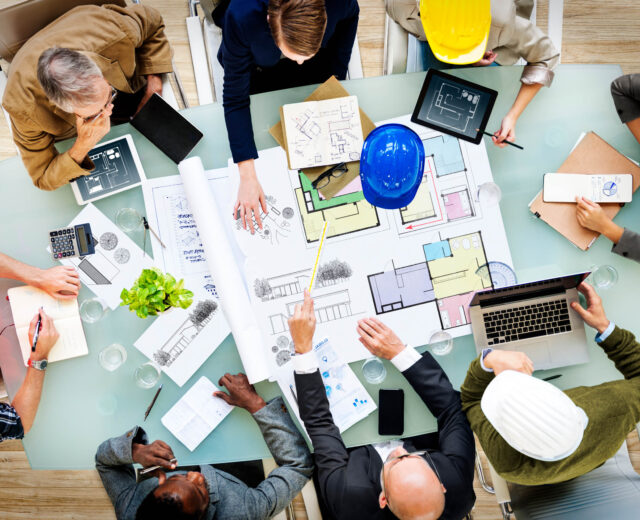 A diverse group of professionals in hard hats and business attire gather around a table, reviewing architectural blueprints and using laptops and tablets.