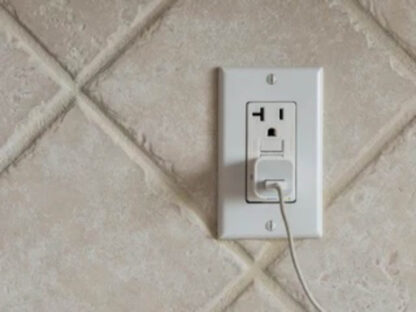 A white electrical outlet with a plug inserted is mounted on a beige tiled wall.