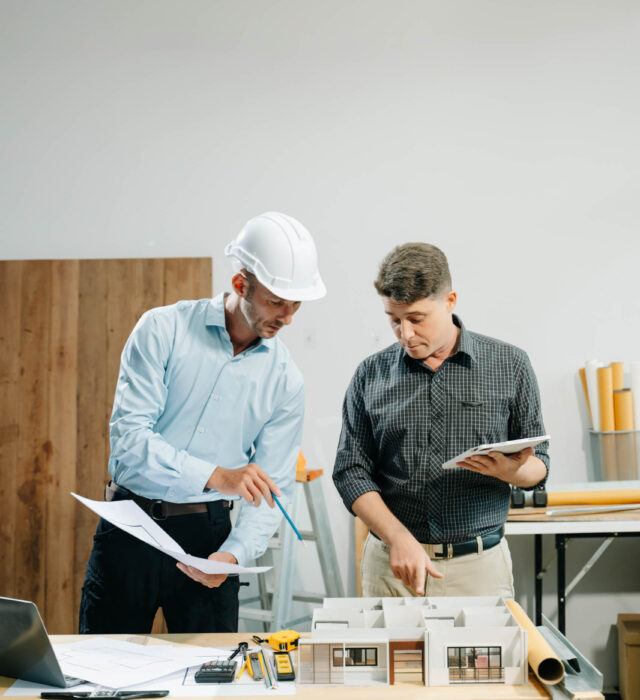 Two men discussing architectural plans at a table with blueprints, a model, and construction tools. One wears a hard hat and holds papers, the other holds a tablet.
