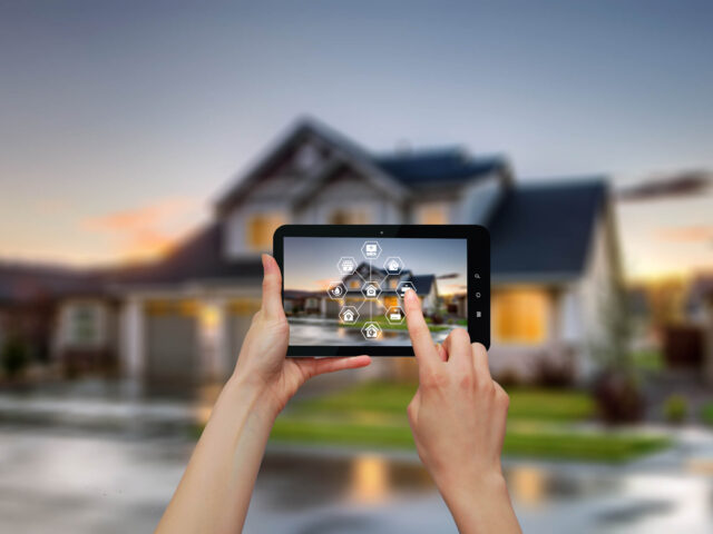 Person using a tablet to control smart home automation features, with a modern house in the background.