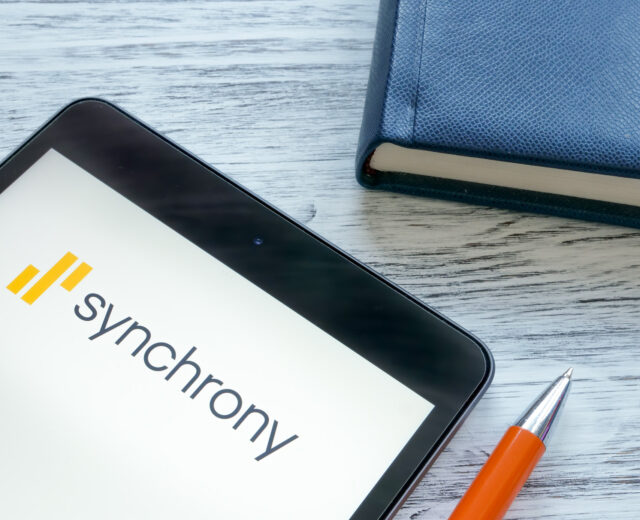 A tablet displaying the Synchrony logo next to a blue notebook and an orange pen on a wooden surface.