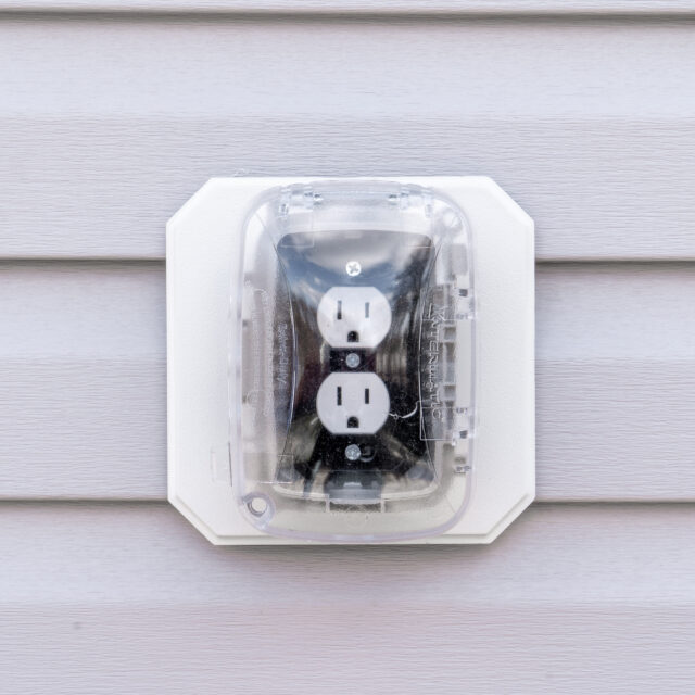Outdoor electrical outlet with cover mounted on a vinyl siding wall.