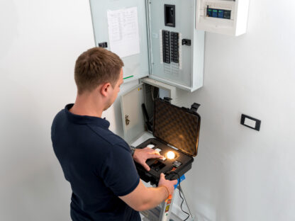 A man stands on a ladder inspecting an electrical panel with a testing device in a case, inside a white room.