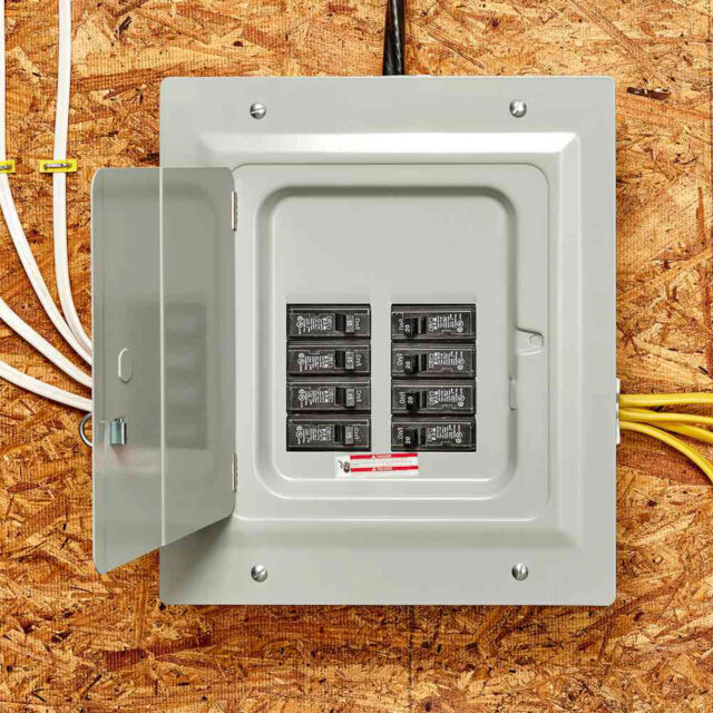 An open electrical panel with circuit breakers mounted on a plywood wall, showing wires entering from the top and right side.