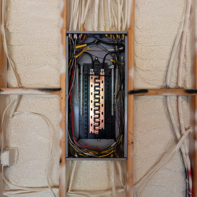 Electrical breaker box with multiple circuit breakers and wires, set within a wall insulated with spray foam.