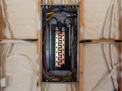An open electrical panel with visible wiring and circuit breakers is set against a wall with insulation foam.