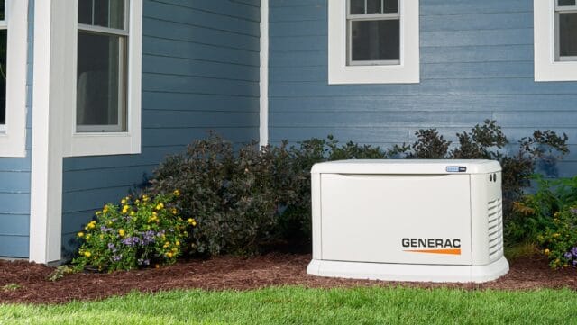 A whole-home Generac generator installed in by the bushes besides a blue house.