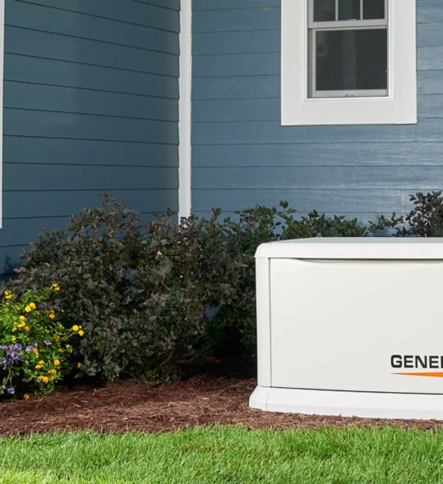 A whole-home Generac generator installed in by the bushes besides a blue house.