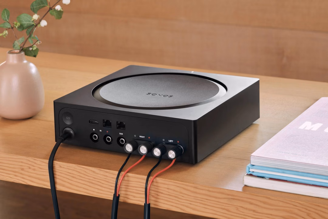 A black Sonos audio amplifier with various cables connected, sitting on a wooden table next to a vase with flowers and a stack of books.