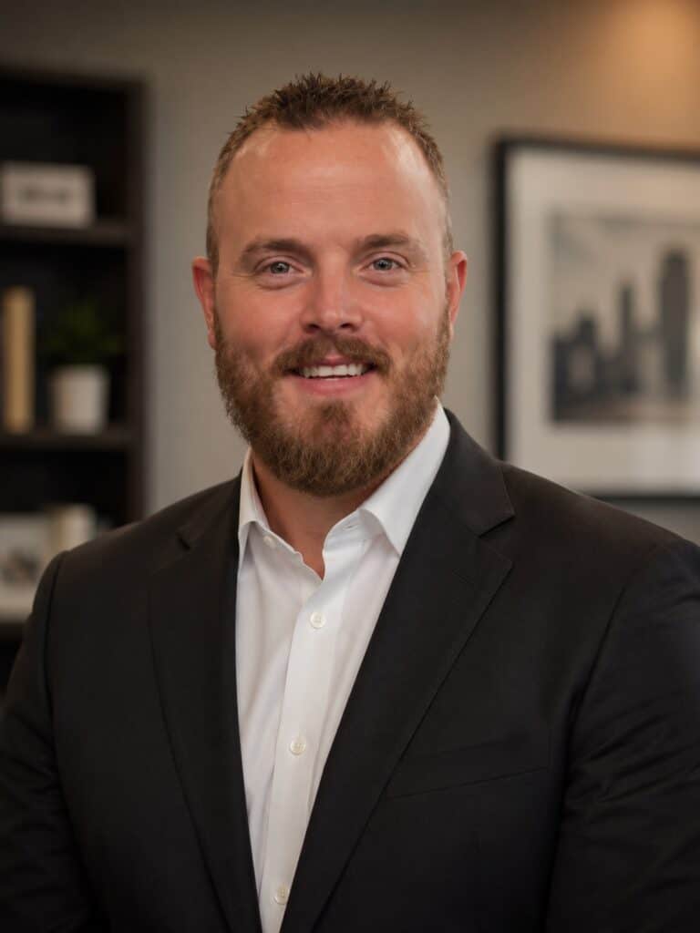 A man with short hair and a beard wearing a dark suit and white shirt stands smiling in an office setting with shelves and framed photo in the background.