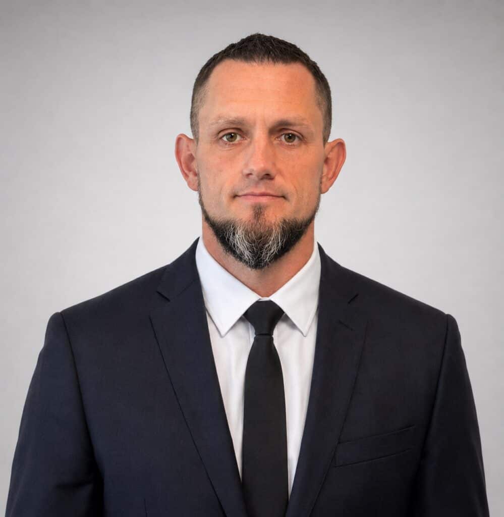 A man with short hair, a beard, and a mustache is wearing a dark suit, white shirt, and black tie, standing against a plain light gray background.